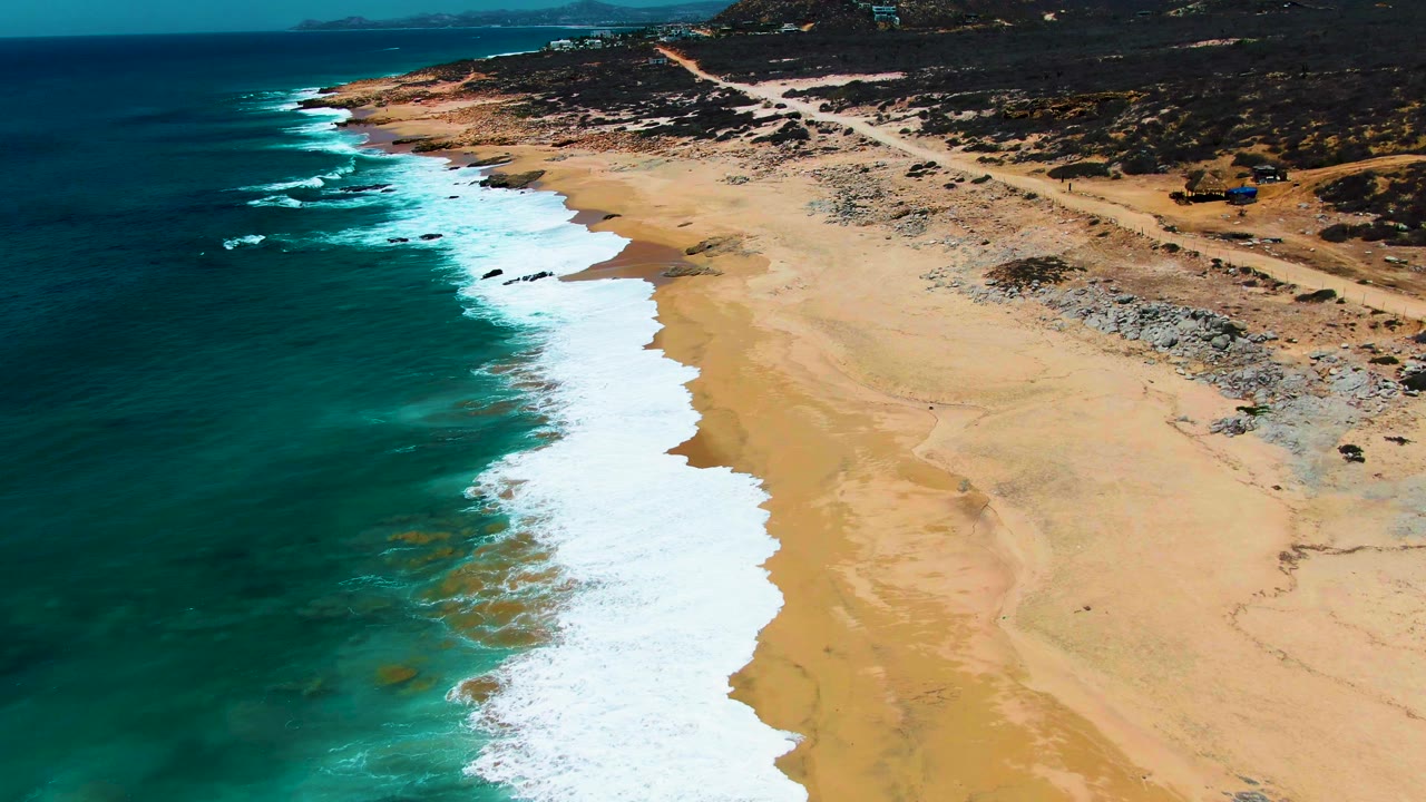 Aerial frame over Buzzards Beach showing a longer band of white foam along the shoreline, dark blue water offshore, open sand on the right, rocky patches near the upper beach, and no visible people.
