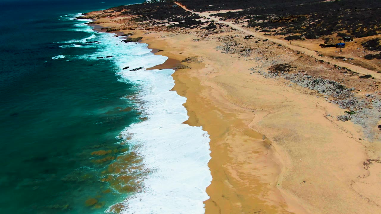 Aerial view at Buzzards Beach with the surf band shifted farther down the frame, sandy beach dominating the right side, rocky sections near the inland edge, coastal scrub beyond the road, and no visible people.