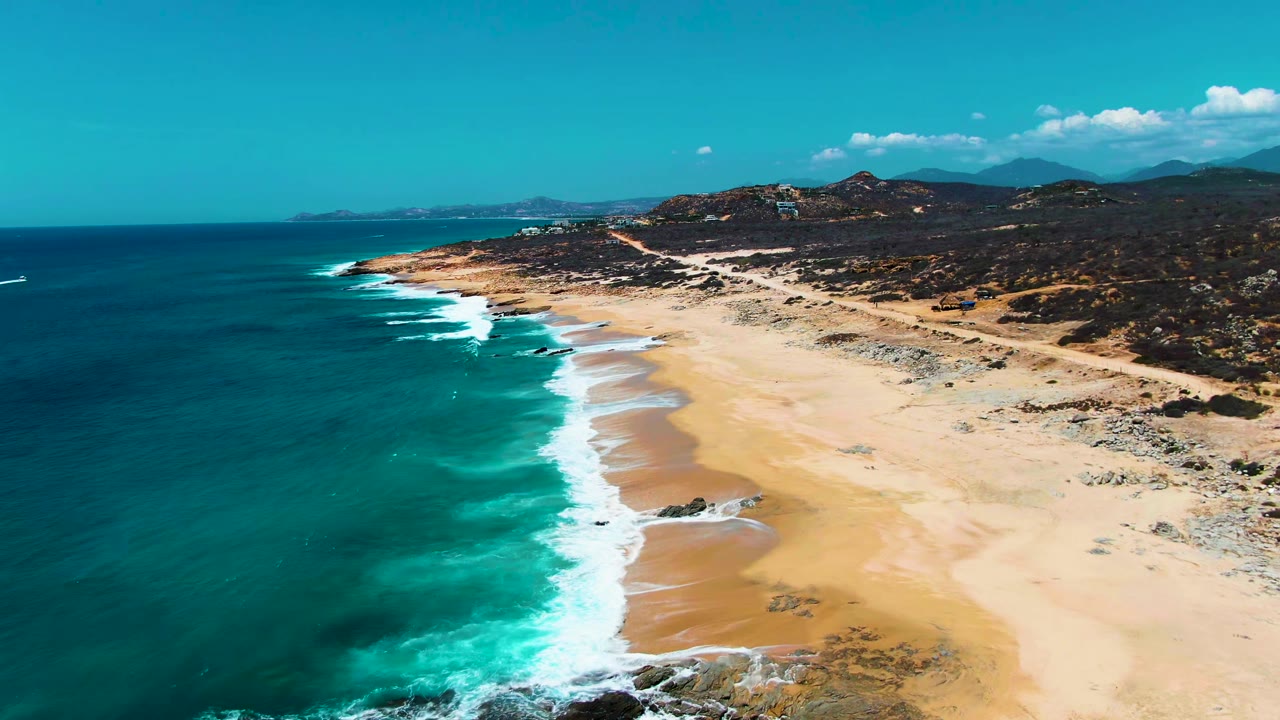 Aerial coastline with turquoise ocean, white surf, tan sand, hills, and road