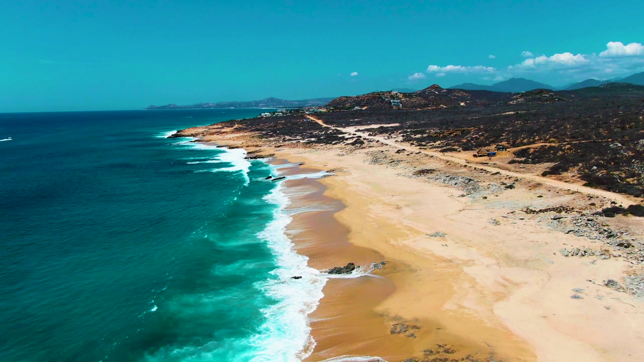 Drone-height view of Buzzards Beach near Punta Buzzards, Mexico, as bands of white foam break along the diagonal shore, with broad sand, scrubby hills, a road, blue sky, and no visible people.
