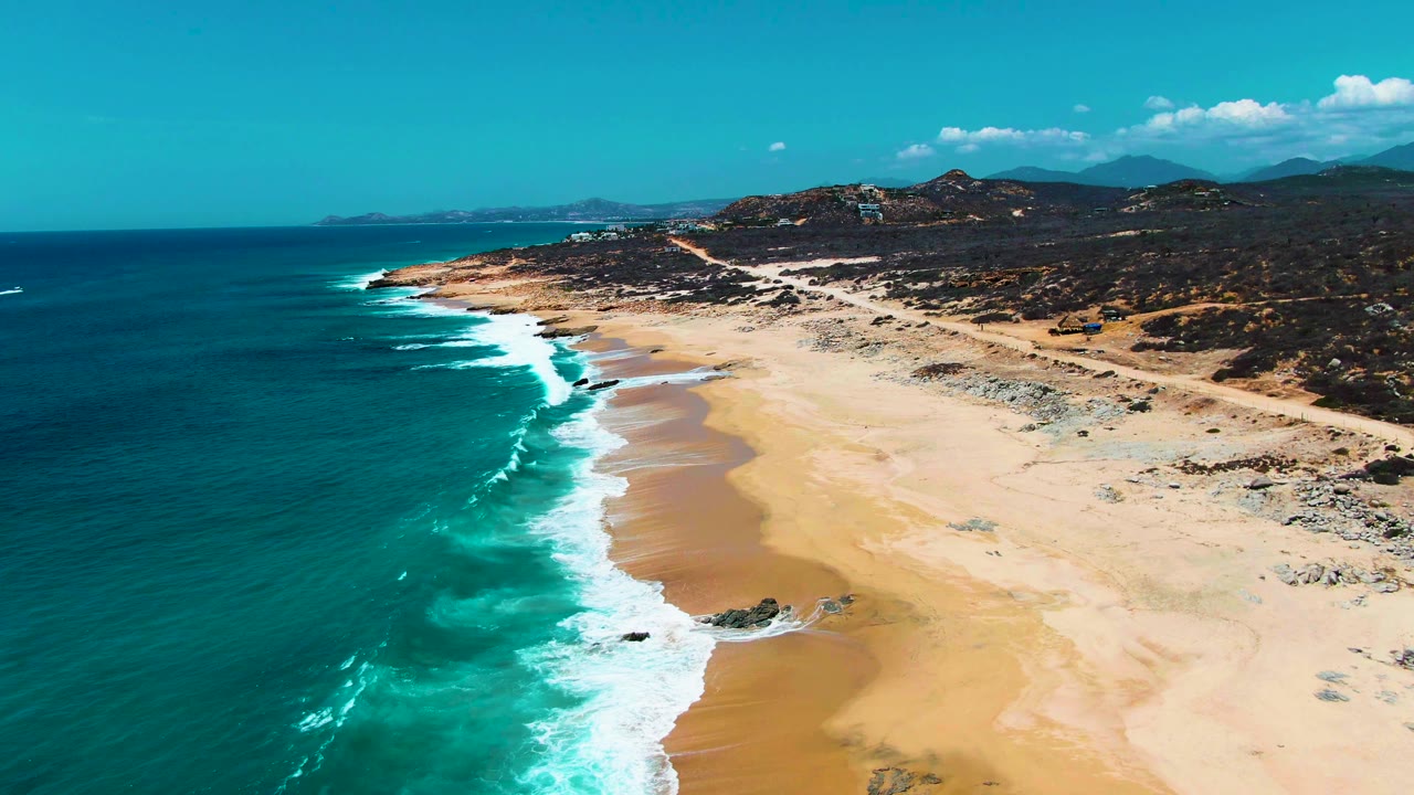 Aerial view at Punta Buzzards with shallow turquoise water near shore, white surf lines sliding onto tan sand, a road above the beach, distant hills, and no visible people.