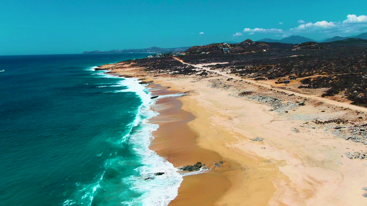 Wide aerial frame over Buzzards Beach, Mexico, with dark blue ocean offshore, white breakers along the beach, scattered rocks on the sand, inland hills and road detail, and no visible people.
