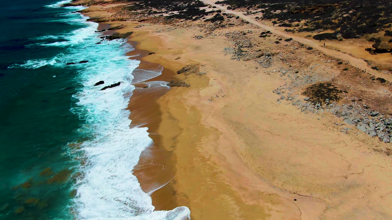 Aerial view over Buzzards Beach in Baja California Sur, with turquoise surf at left, broad tan sand below, rocky scrubland inland, and no visible people.