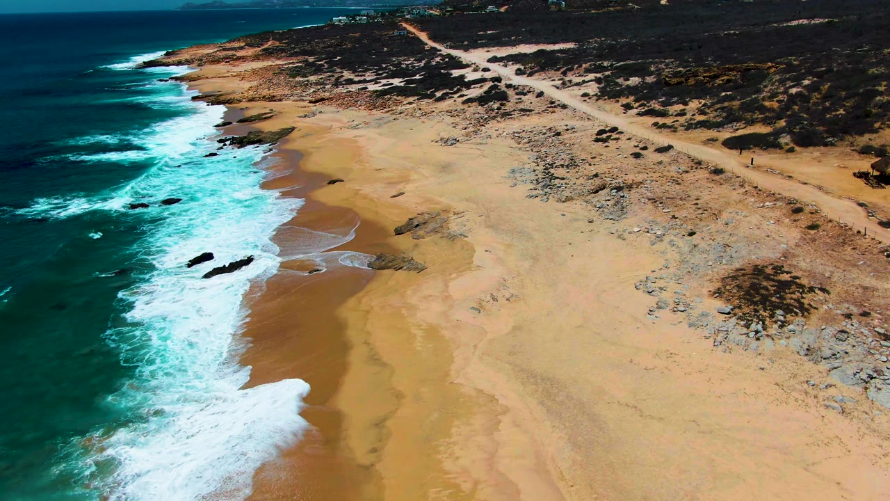 Aerial frame at Buzzards Beach showing a longer stretch of breaking waves, wet brown sand, scattered rocks, inland scrub, a parallel dirt road, and no visible people.