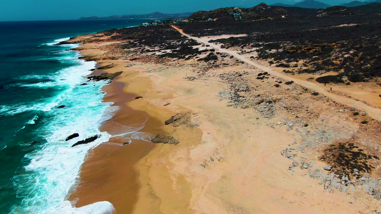 Oblique aerial view along Buzzards Beach, with blue-green ocean on the left, white surf lines, tan sand, dark scrub-covered hills inland, and no visible people.