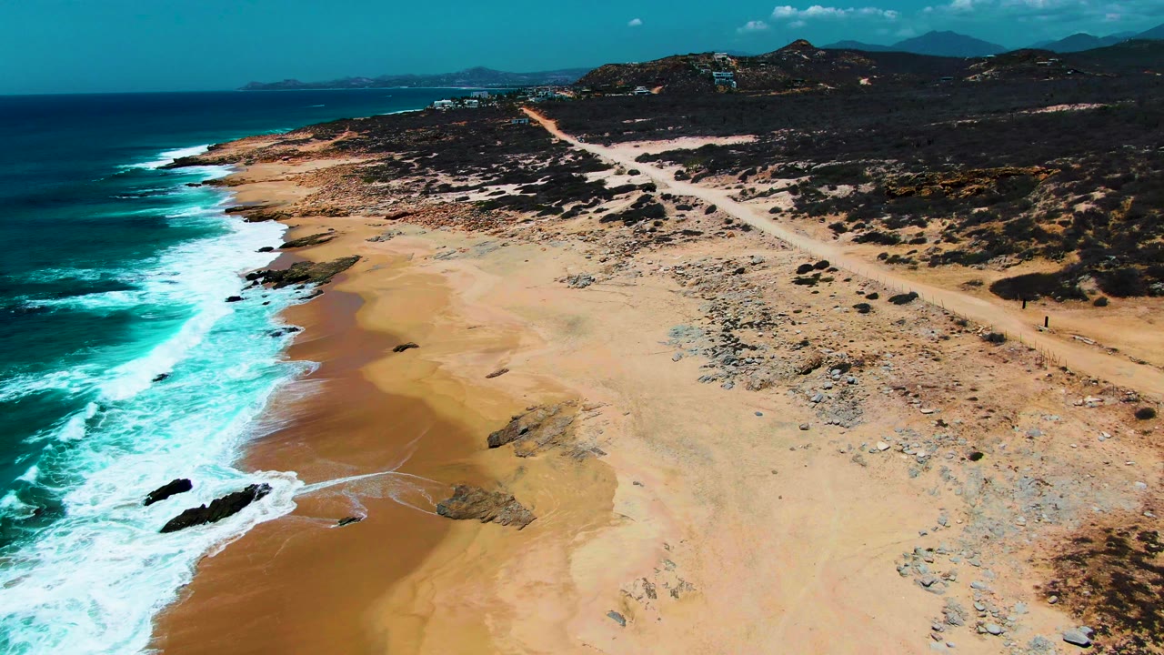 Aerial coastline with waves hitting sand, rocky beach, dirt track, and low hills.
