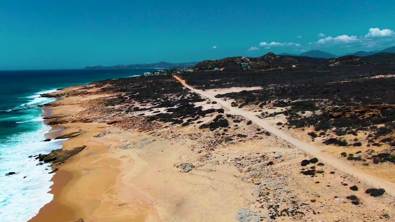 Aerial frame over Buzzards Beach in Baja California Sur, with ocean surf on the left, pale sand below, a dirt road through scrubland, distant hills, and no visible people.