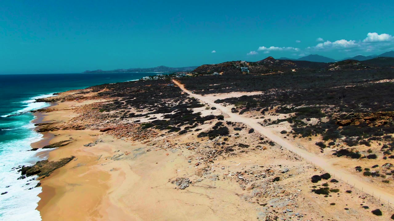 Aerial view of Punta Buzzards coastline with white waves reaching the sandy shore, a diagonal dirt road across rocky scrub, low hills beyond, and no visible people.