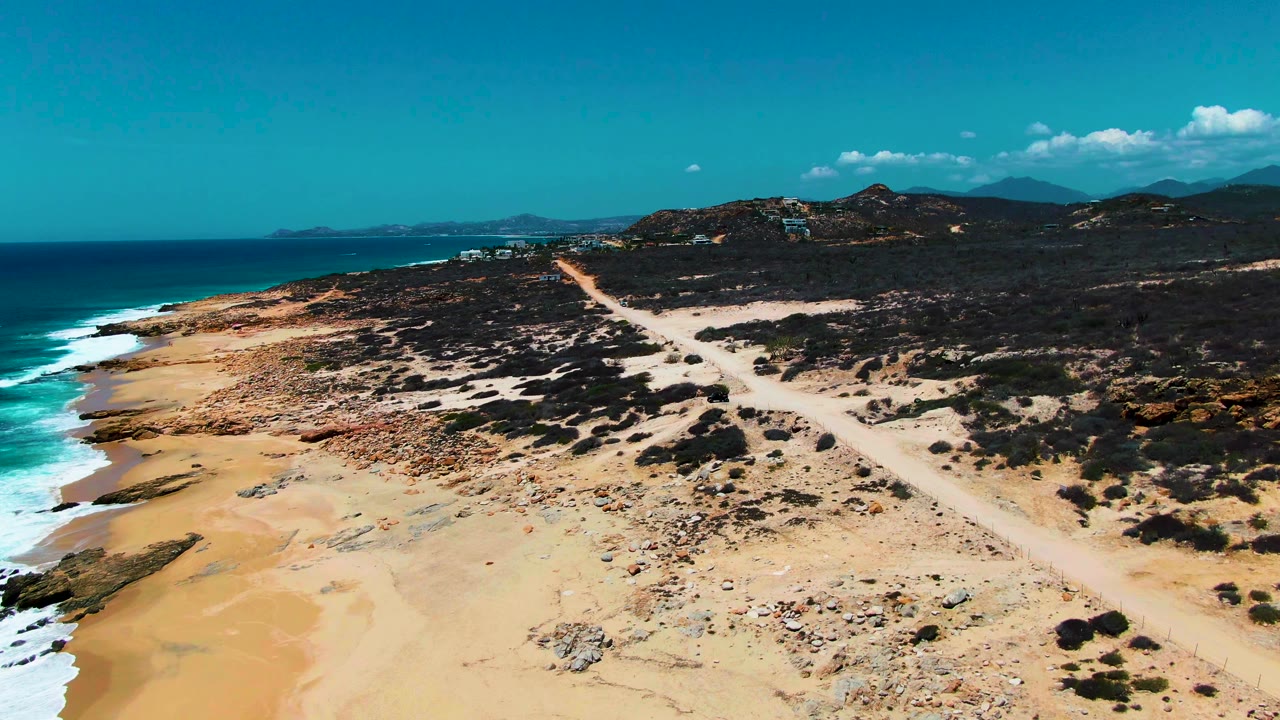 Drone-style frame of the East Cape beach, where blue ocean and white surf border tan sand while a pale road cuts through dark coastal vegetation with no visible people.