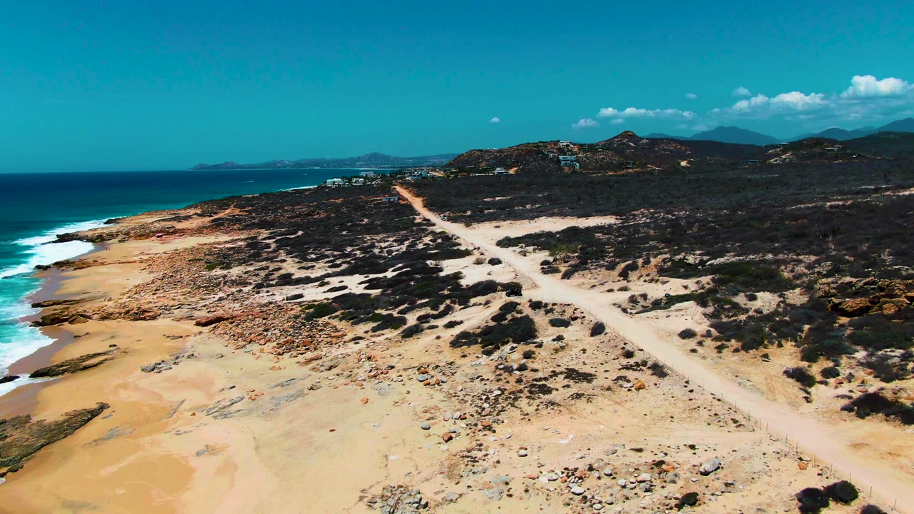 Aerial beach landscape with ocean surf, rocky sand, scrub, and a dirt track.