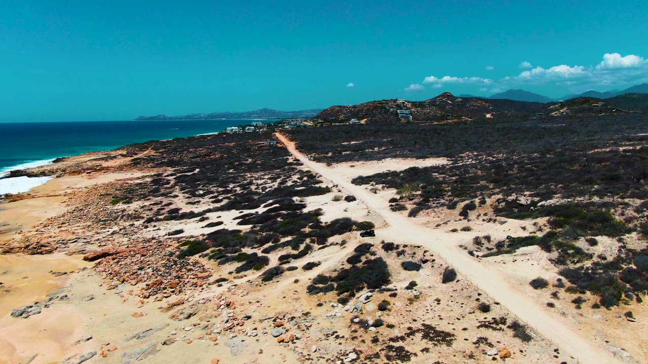 At Punta Buzzards in Baja California Sur, an elevated view shows a pale dirt road cutting through sandy scrub toward low hills, with blue ocean on the left and no visible people.