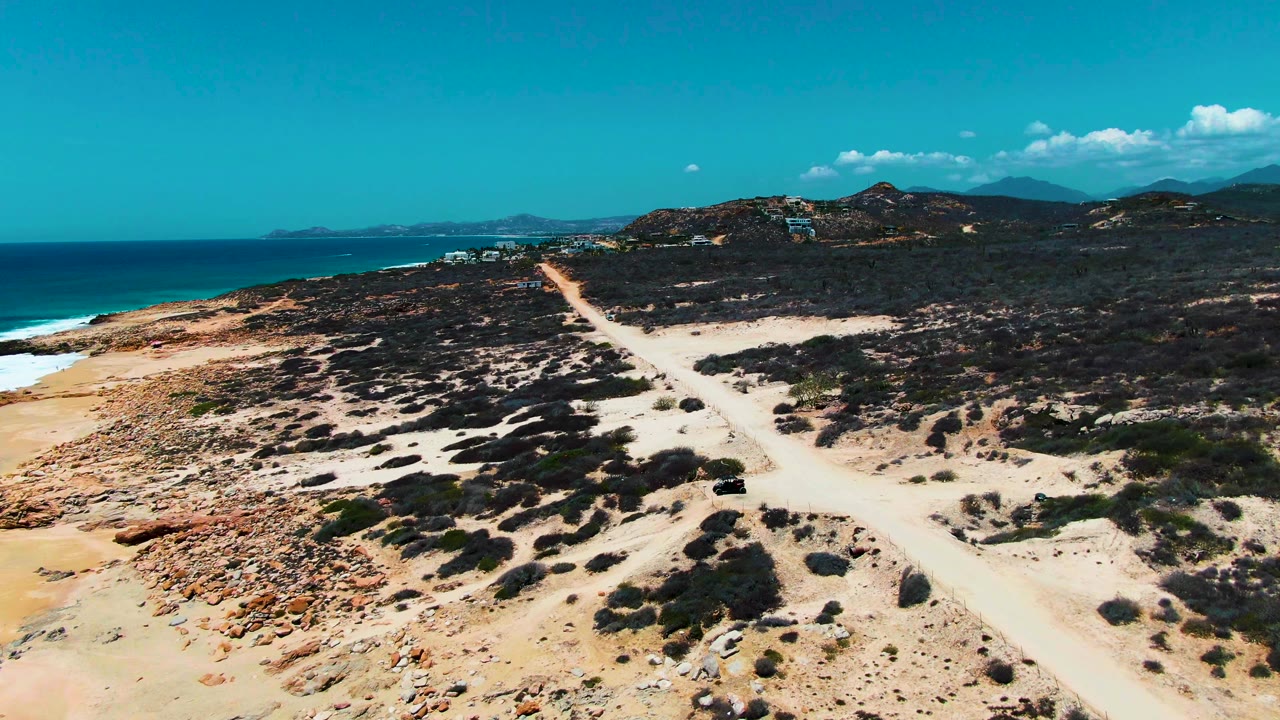 Aerial coastal road scene with ocean, sand, scrub, hills, and a small dark vehicle.