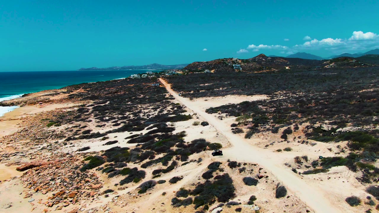 At 0:02, the black vehicle is lower in the frame on the pale road, surrounded by sand and dark scrub, with the turquoise ocean, low hills, and clouded blue sky in the background, and no visible people.