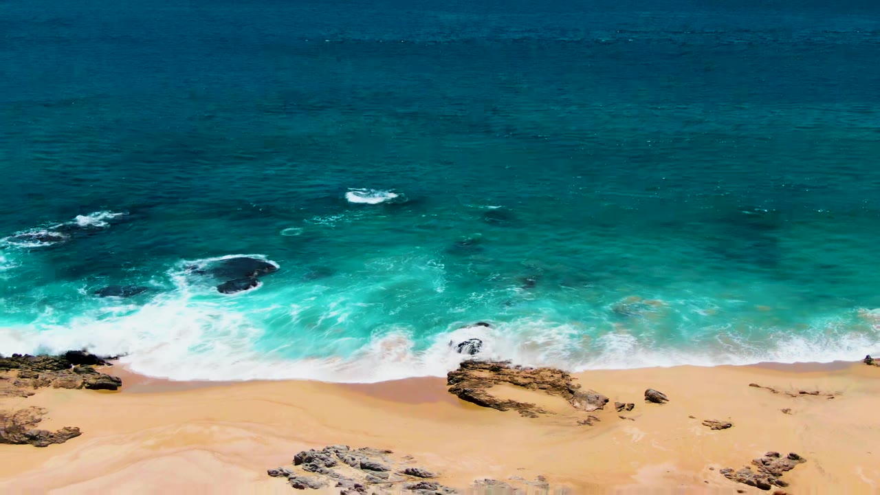 Aerial view of turquoise waves reaching a sandy beach with rocky outcrops.