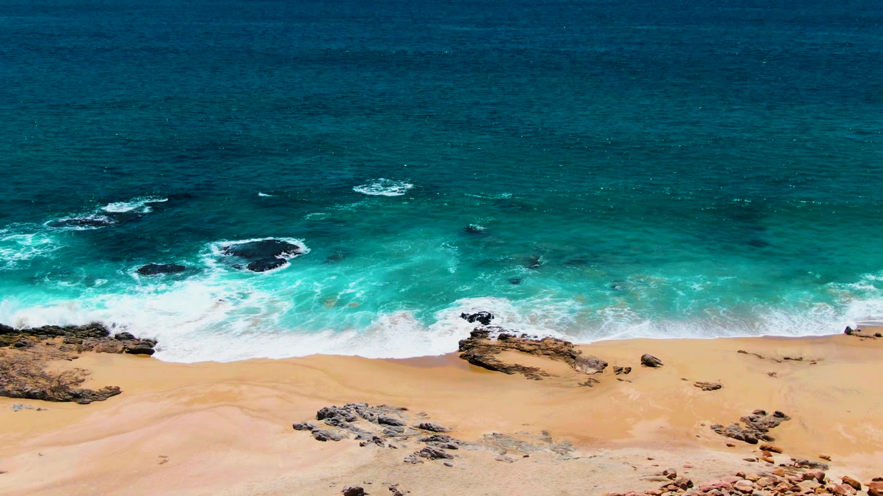 Aerial frame from Buzzards Beach showing white foam spreading around several shoreline rock ledges, with clear turquoise water and no visible people.