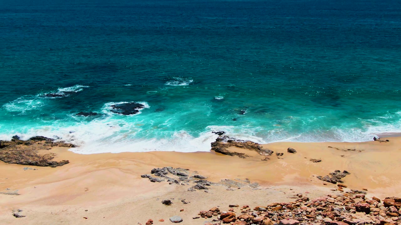 Overhead Buzzards Beach view with a broad turquoise band of surf, pale sand below, scattered rock outcrops, and no visible people.