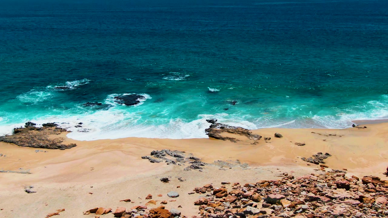 Buzzards Beach aerial still with white water advancing over tan sand, small rock shelves in the surf, reddish stones at the bottom edge, and no visible people.