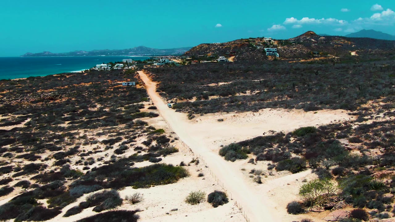 At 0:00 in Punta Buzzards, Baja California Sur, a sandy dirt road runs through dry scrub with turquoise ocean on the left, scattered buildings ahead, low hills beyond, and no visible people.