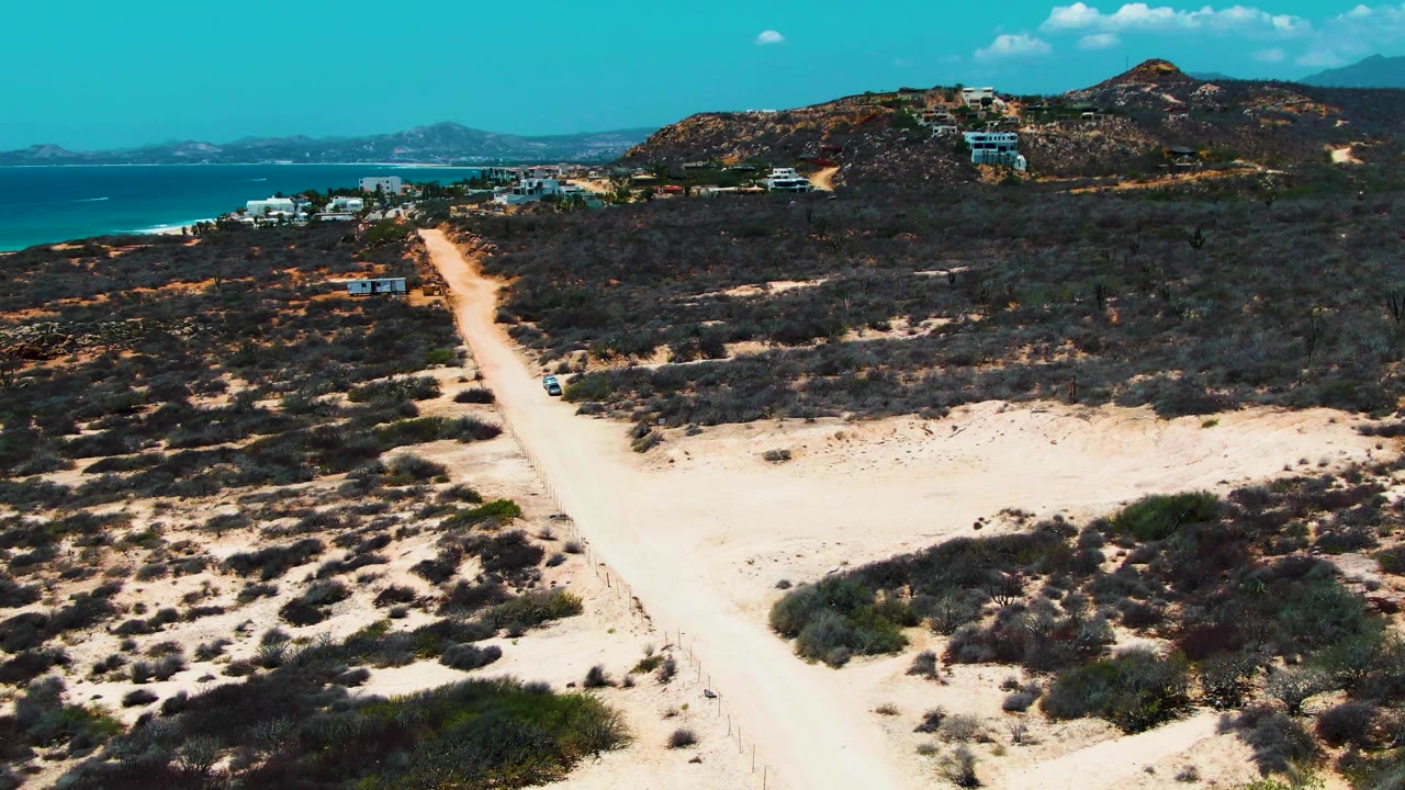 At 0:02, the car is farther along the sandy track through sparse desert vegetation, with the Baja California Sur coast, hillside homes or buildings, and clear blue sky visible, and no visible people.