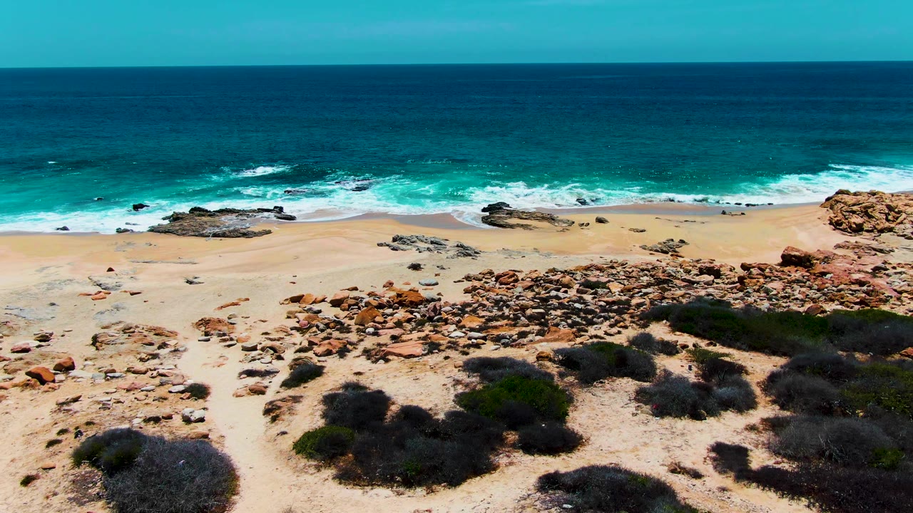Aerial frame over Buzzards Beach in Baja California Sur showing a broad tan sand strip, scattered reddish rocks, turquoise surf, and no visible people.