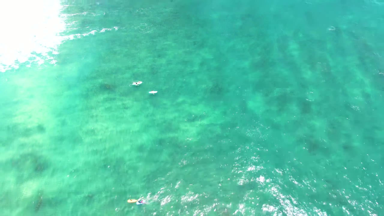 Overhead view of clear turquoise water at Punta Buzzards with bright surf along the left edge and a few small swimmers or floating figures near the center and lower left.