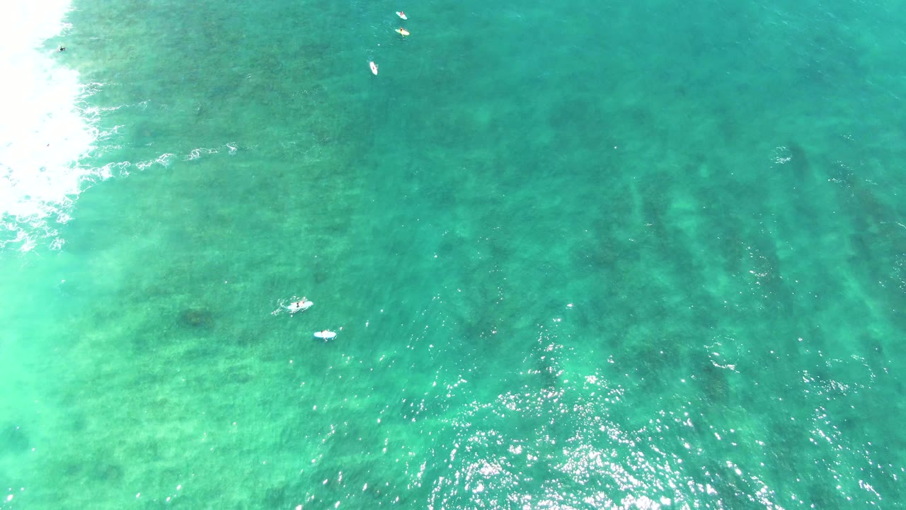 Overhead Punta Buzzards ocean frame with bright surf at the far left, dark underwater streaks, and several tiny swimmers or boards near the top and midframe.