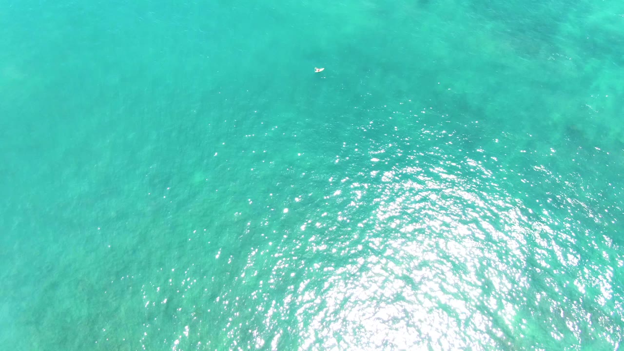 Top-down view of clear turquoise ocean at Punta Buzzards with a few tiny swimmers across the frame, a strong patch of sun shimmer to the right, and no shoreline visible.