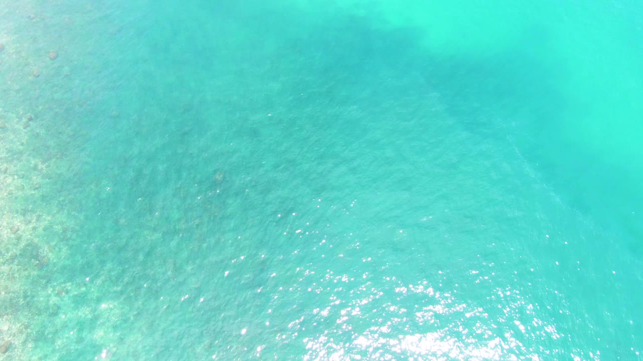 A bright overhead view of Buzzards Beach water shows faint seabed texture under turquoise ripples, with stronger sunlight reflection near the lower center and no visible people.