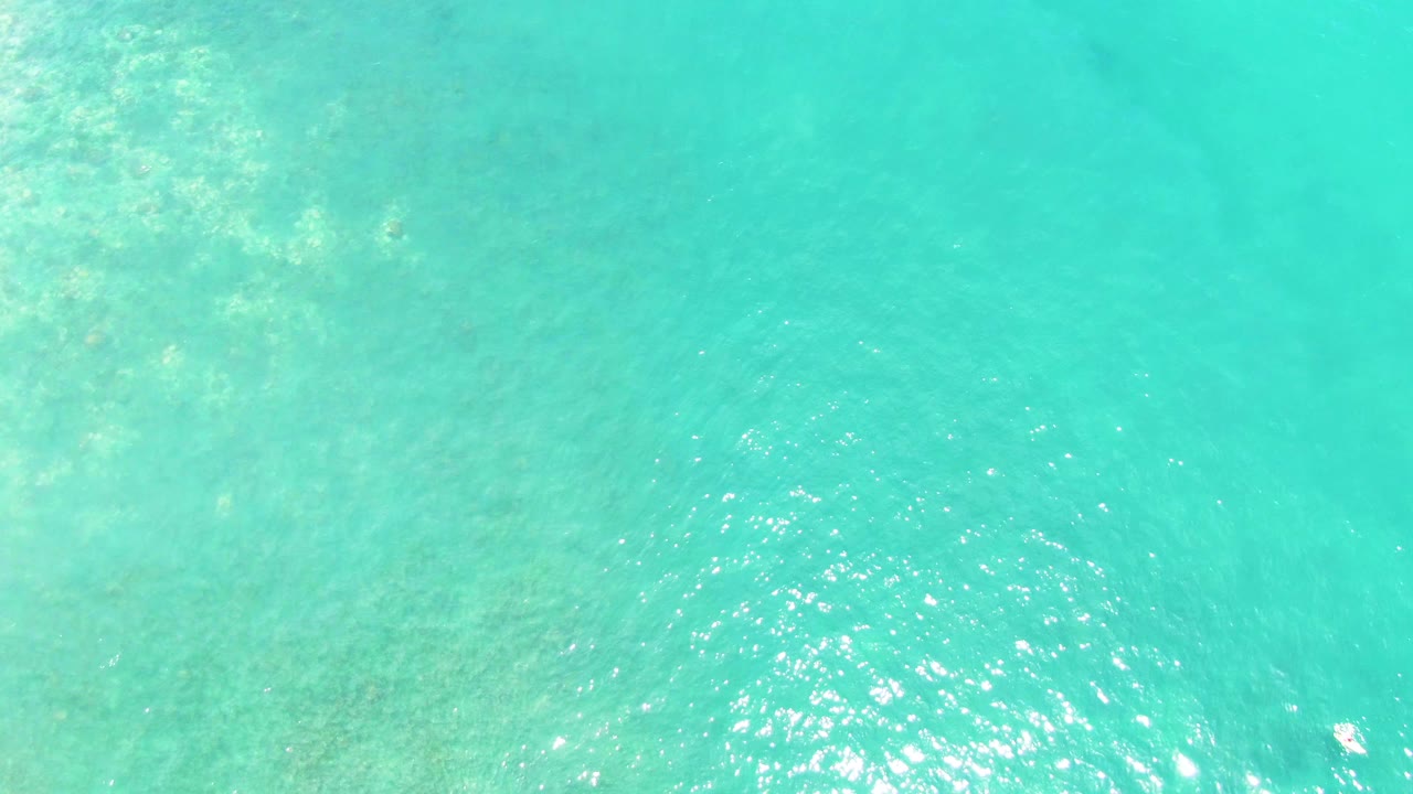 Overhead view of clear turquoise water at Buzzards Beach, Baja California Sur, with mottled seabed texture in the upper left, bright surface reflections near the lower center, and no visible people.