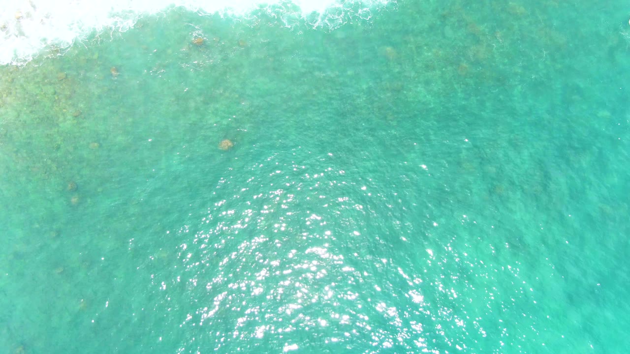 Overhead view of turquoise shallow water at Punta Buzzards, with white wave foam entering along the upper edge, sun glints on ripples, and no visible people.