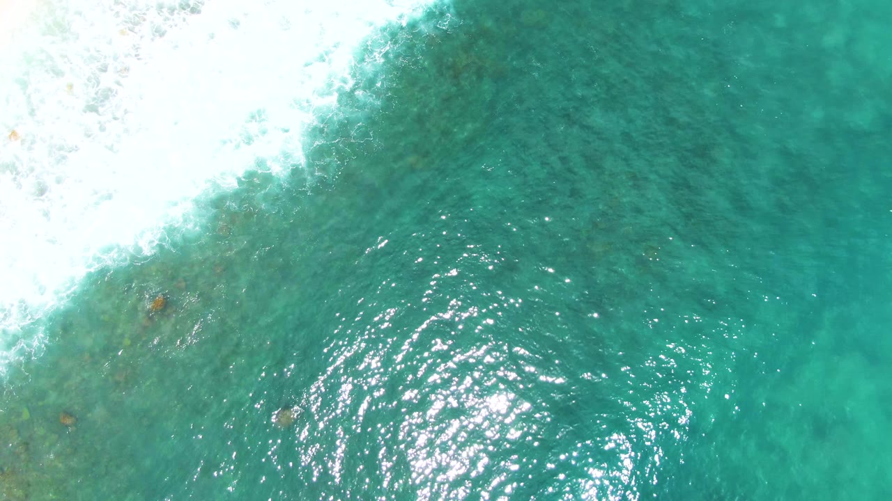 Aerial frame at Punta Buzzards showing white surf sweeping diagonally from the upper left over darker turquoise water, with ripples and no visible people.