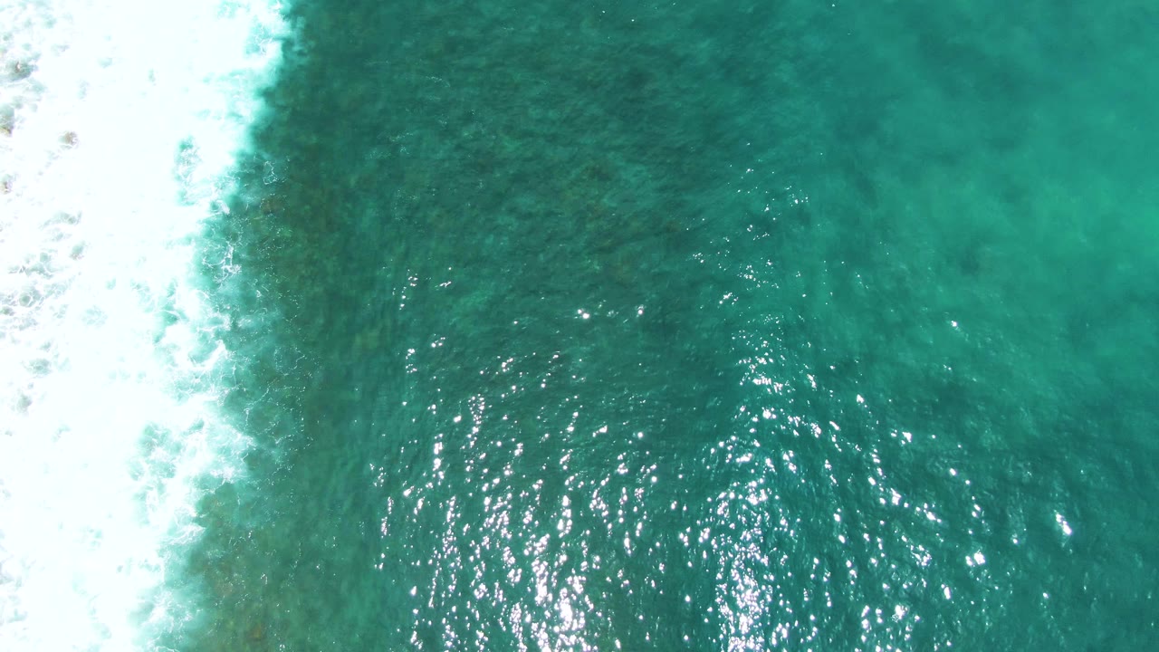 Overhead Punta Buzzards frame with white foam pushing in from the left edge, darker green-blue water filling most of the view, bright reflected highlights, and no visible people.