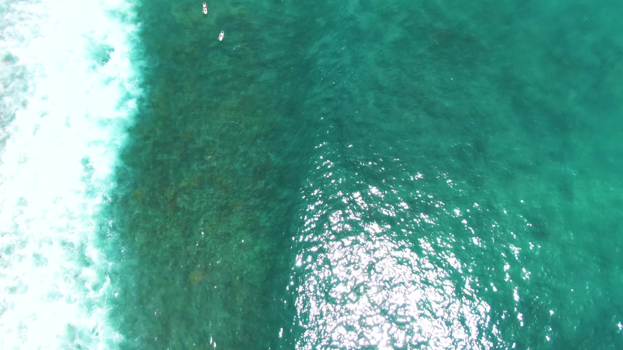 At Punta Buzzards, an overhead aerial frame shows turquoise ocean water, white surf foam along the left side, sun glare on ripples, and small swimmers near the top edge.