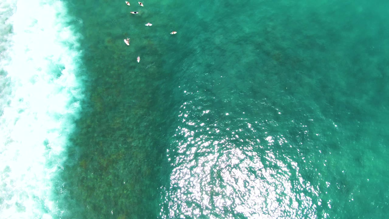 Aerial turquoise water with breaking surf on the left and several swimmers near the upper center.