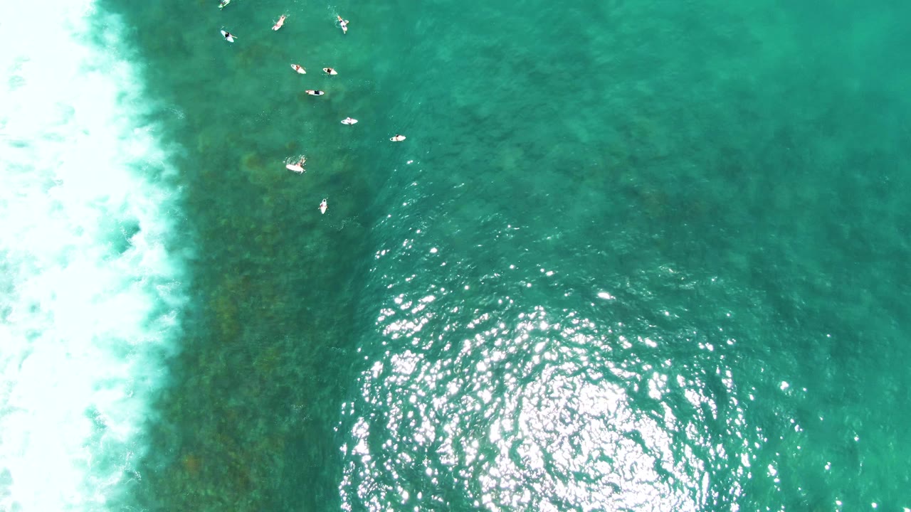 At Punta Buzzards, swimmers sit near the top of the aerial frame while a large bright sun reflection spreads across the clear turquoise water and surf foam remains at the left edge.