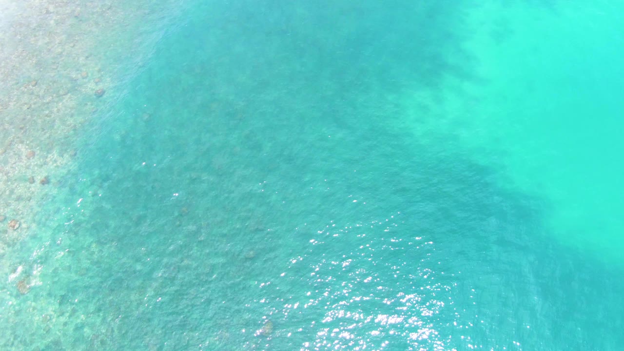Overhead view of turquoise shallow ocean water at Punta Buzzards, with faint seabed texture and small sunlight glints, no visible people.