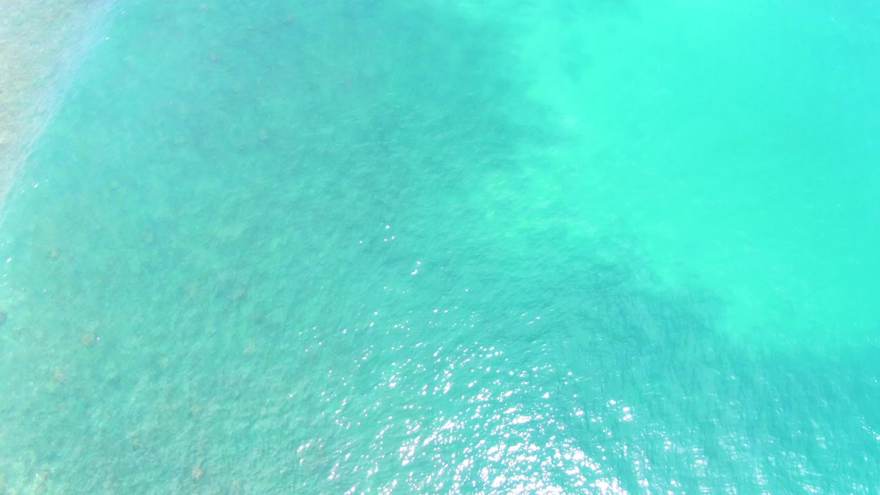 Top-down turquoise sea at Punta Buzzards shows darker underwater patches toward the upper left and scattered sun flecks across the surface, no visible people.