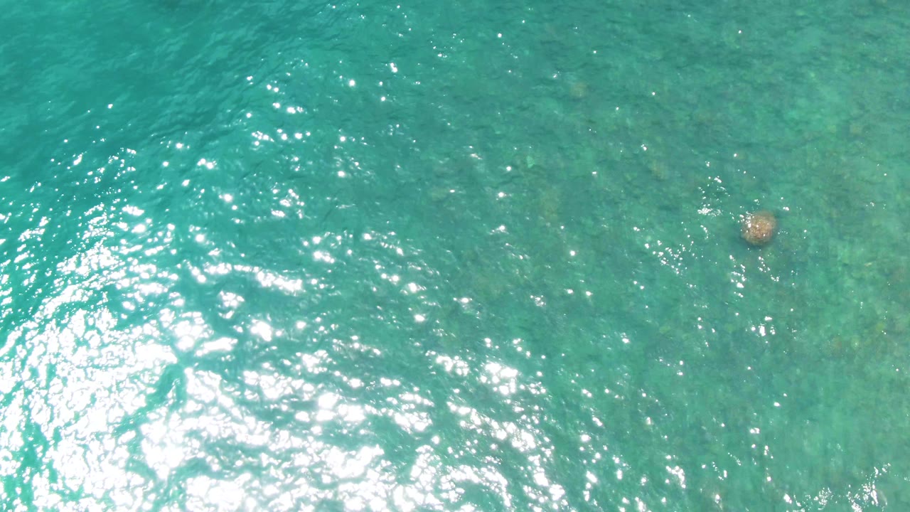 Top-down view of turquoise water at Punta Buzzards, Baja California Sur, with bright surface glare and a submerged rock near the upper center, no visible people.