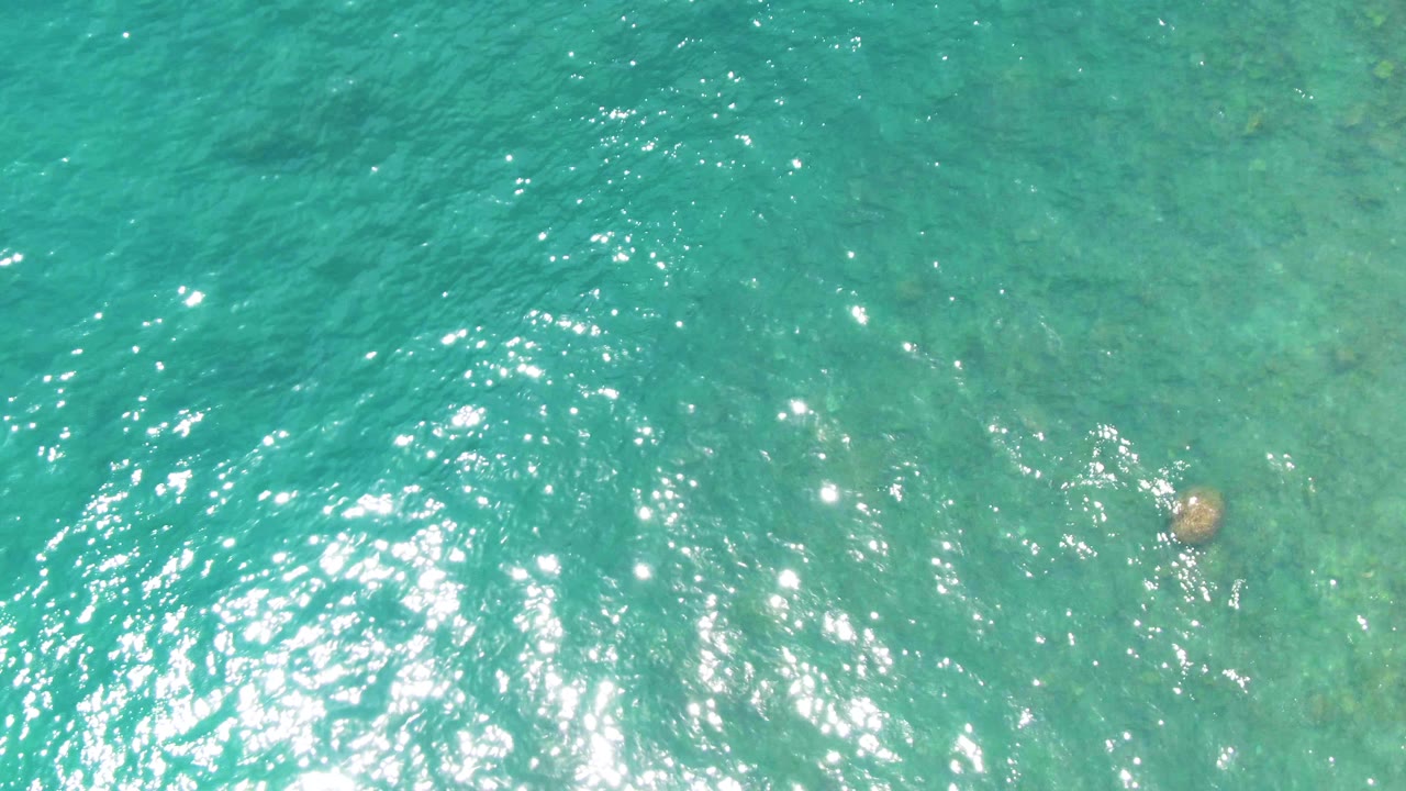Overhead frame of clear blue-green ocean at Punta Buzzards, Baja California Sur, with a single submerged rock and fine ripples across the surface, no visible people.