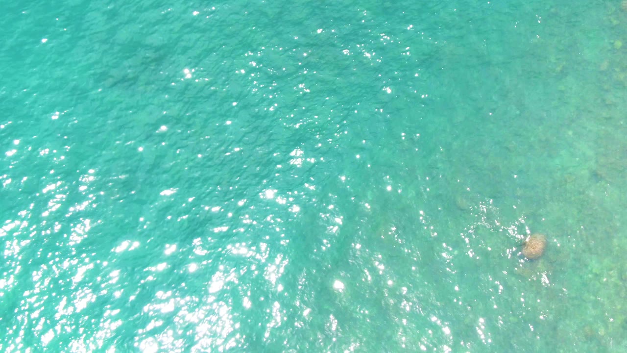 Top-down turquoise water at Punta Buzzards, Baja California Sur, with soft sun sparkle, faint seabed color changes, and a submerged rock toward the right side, no visible people.