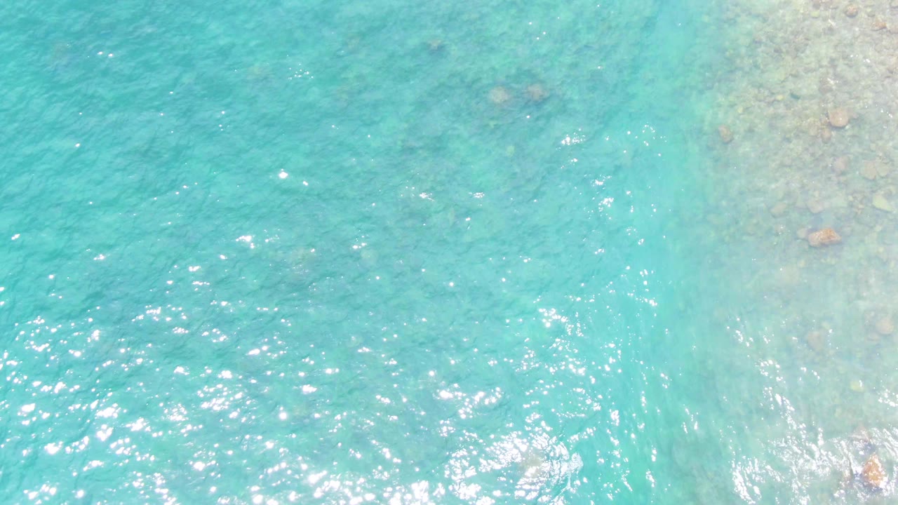 Top-down view of turquoise water over submerged rocks at Punta Buzzards in Baja California Sur, with sun glints on the rippled surface and no visible people.