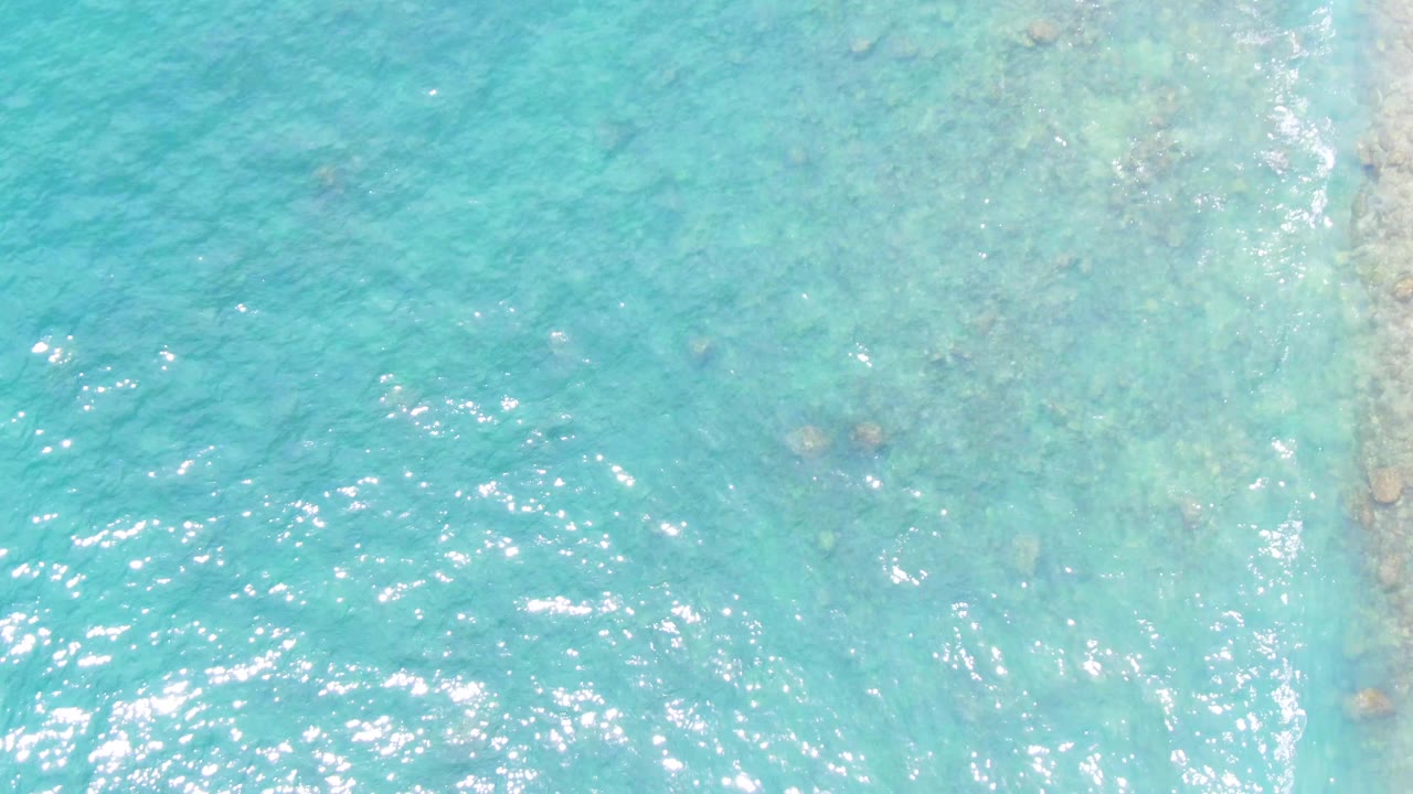 Turquoise shallow water at Punta Buzzards with a pale rocky shoreline area near the right edge, visible seabed texture, and no visible people.