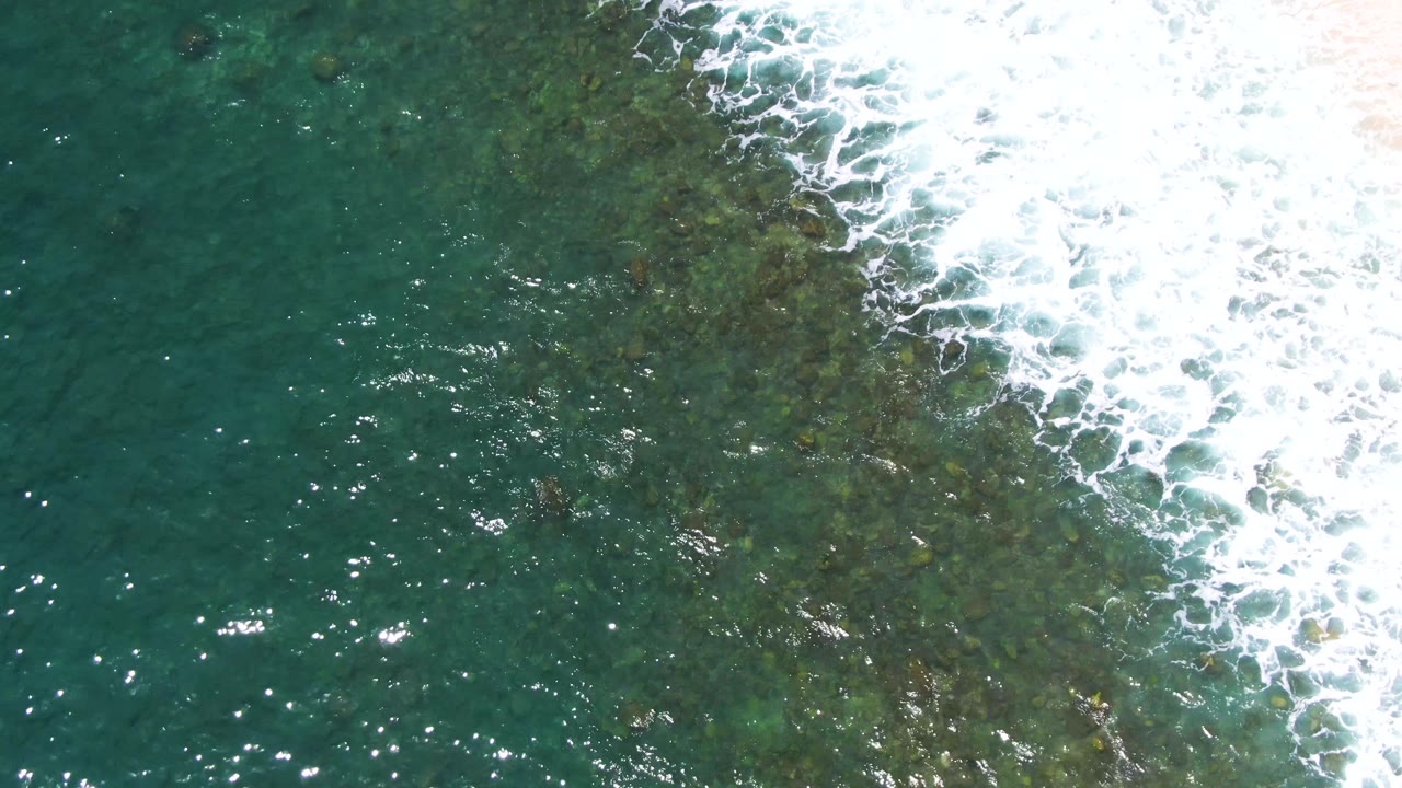 A foamy wave edge crosses the upper right of the overhead frame at Buzzards Beach, with green water and mottled seafloor texture below, and no visible people.