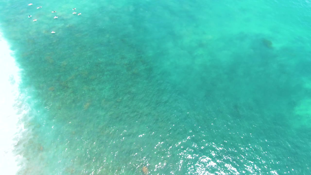 Overhead view at Buzzards Beach, East Cape, Baja California Sur, showing turquoise water, distant swimmers near the top of frame, darker seabed patches, and white surf along the left edge.
