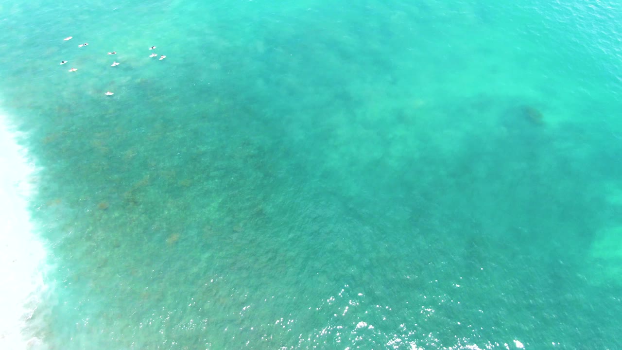 Turquoise ocean from above with small swimmers near the top and surf on the left.