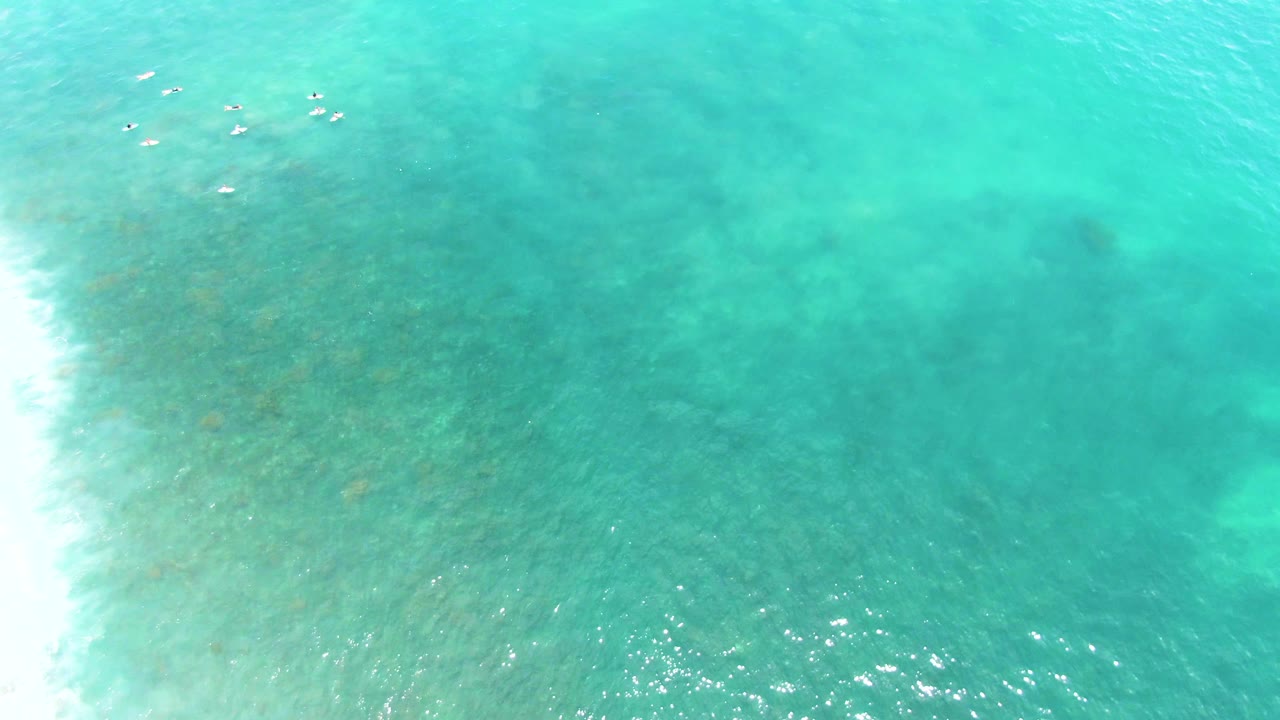 At Buzzards Beach, East Cape, Baja California Sur, the overhead view shows distant swimmers near the top, a bright band of surf at left, and rippled turquoise water with darker areas below the surface.
