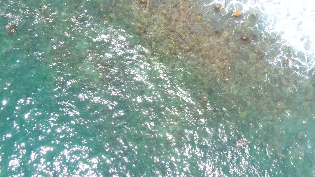 Top-down view of turquoise water over a rocky seabed at Punta Buzzards, Baja California Sur, with white surf foam at the upper right and no visible people.