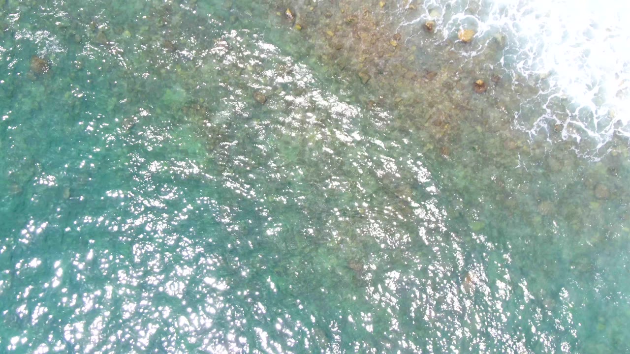 Aerial frame of clear blue-green ocean at Punta Buzzards with bright surface reflections, submerged rocky patches, and no visible people.