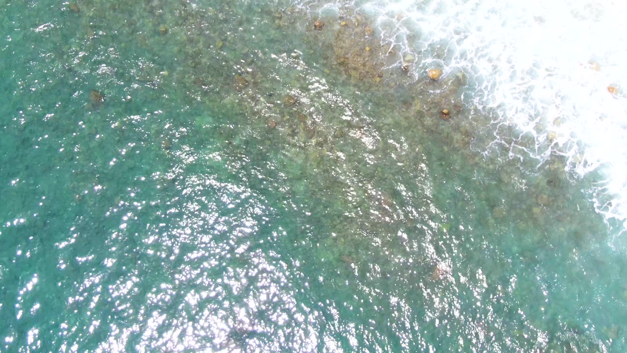 Top-down coastal water at Punta Buzzards with white foam crossing the upper right, mottled reef texture below the surface, and no visible people.