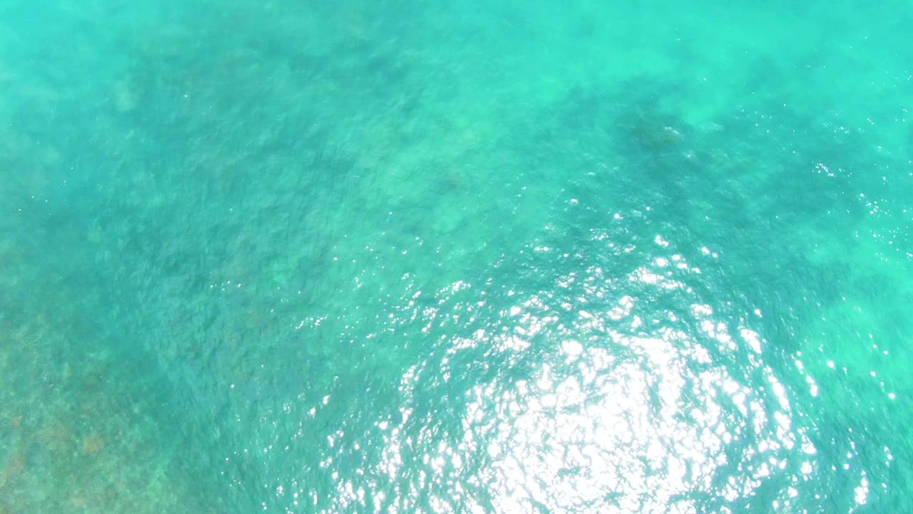 Top-down view of turquoise ocean water at Punta Buzzards, Baja California Sur, with a bright sun reflection and shallow seabed texture, no visible people.